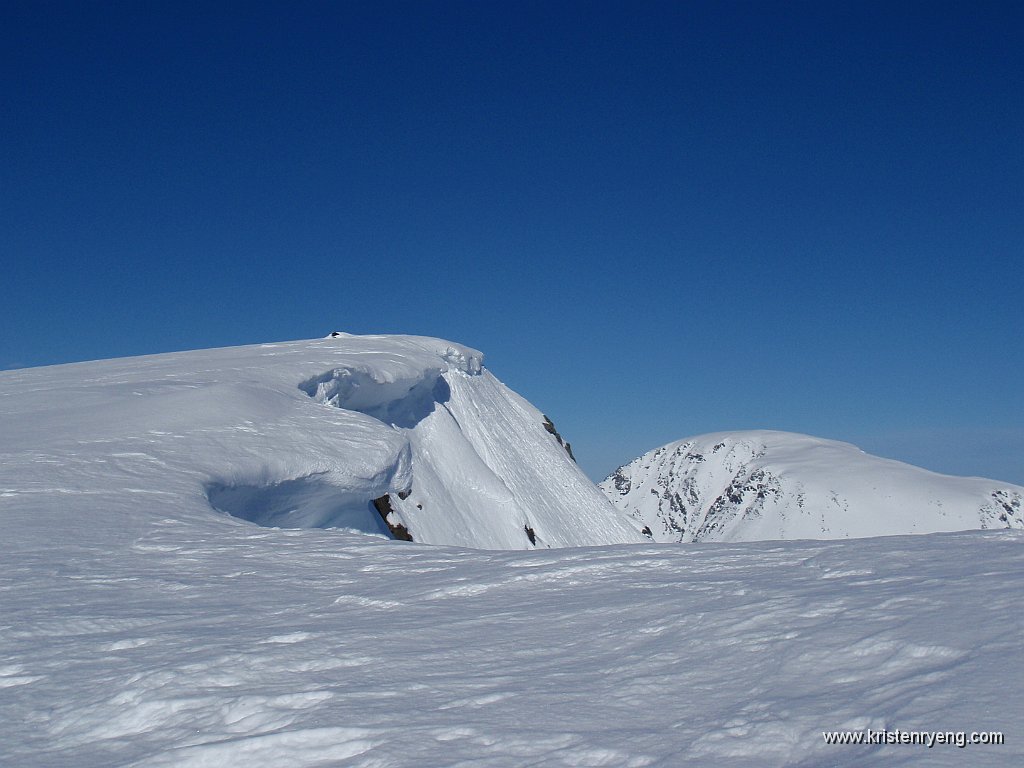P4030426.JPG - Store skavler henger utenfor toppen. En godt nedsnødd varde ses såvidt på toppen. Bak til høyre ses Nordfjellet (1000 moh).